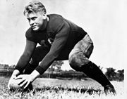 A uniformed but helmetless American Football player is shown on a football field. He is in a ready position, with legs in a wide stance and both hands on a football in front of him.