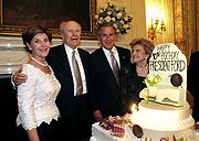 Two men in suits are flanked by two women in formal dresses, standing beside a large birthday cake with lit candles and flowers. The cake is decorated with the text 