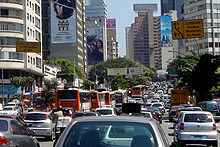 Looking down a busy road, which is banked on both sides by tall buildings, some of which are covered in advertisement billboards