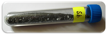 A clear vial containing small chunks of a slightly lustrous black solid, labeled