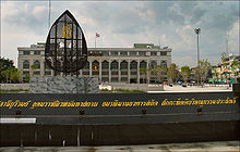 A granite sign with a long name in Thai script, and a building in the background