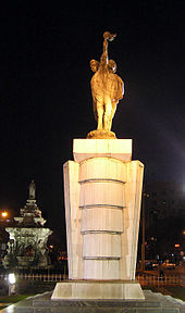 A stone statue of torch-bearers as seen at night. A fountain with a white base is in the background