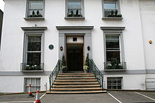 A flight of stone steps leads from an asphalt car park up to the main entrance of a white two-story building. The ground floor has two sash windows, the first floor has three shorter sash windows. Two more windows are visible at basement level. The decorative stonework around the doors and windows is painted grey.