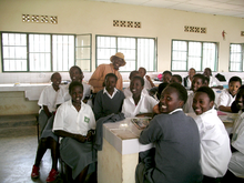 Photograph depicting 18 female school children with white and grey uniforms, and a male teacher with orange shirt and hat, with white tiled sink and surfaces and windows in background