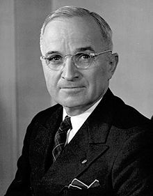 A middle-aged Caucasian male wearing a dark business suit and wireframe glasses is depicted smilingly pensively at the camera in a black-and-white photo.